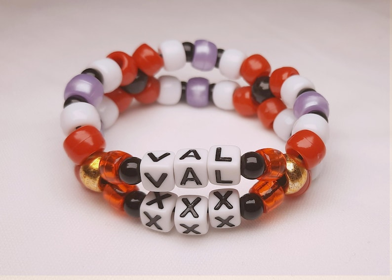 Peut inclure: Deux bracelets de perles. Le bracelet sup&eacute;rieur pr&eacute;sente des perles rouges, blanches et violettes. Le bracelet inf&eacute;rieur comporte des perles de lettres blanches &eacute;pelant "VAL" et "XXX". Les bracelets sont faits de petites perles rondes de diff&eacute;rentes couleurs.