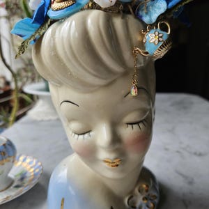 May include: A white ceramic bust of a woman with blue and gold accents. The bust is decorated with a floral arrangement of blue and white flowers, and a small blue teapot charm hangs from the woman's ear.