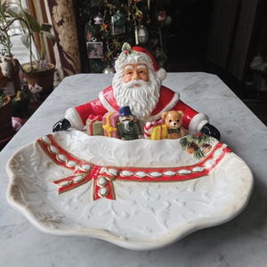 May include: A ceramic Christmas serving platter featuring Santa Claus with gifts and a teddy bear. The white platter has a red ribbon design and embossed floral patterns. The Santa Claus figure is in red and white.