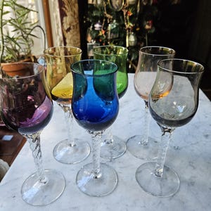 May include: A collection of six colorful wine glasses. Each glass features a different hue, including purple, yellow, blue, green, pink, and gray. The stems have a twisted design, and the bases are clear and circular.