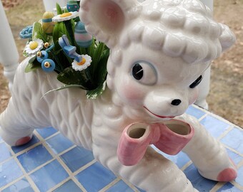 May include: White ceramic lamb planter with a pink bow, holding a bouquet of artificial flowers with blue and yellow Easter eggs.