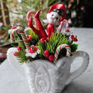 May include: A white milk glass pitcher with a festive Christmas arrangement. The arrangement includes a red and white candy cane, red berries, green pine sprigs, and a red elf figurine.