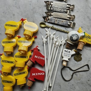 May include: A collection of vintage barware items, including bottle openers, bottle stoppers, and cocktail stirrers. The bottle openers are silver-toned, and the stoppers are yellow and red. Some items have brand names like "Fleischmann's" and "Ballantine" printed on them.