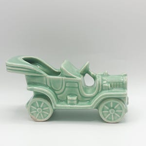 May include: A light green ceramic antique car figurine. The vintage vehicle features detailed wheels, a steering wheel, and a classic open-top design. The decorative piece is a charming addition to any collection or display.