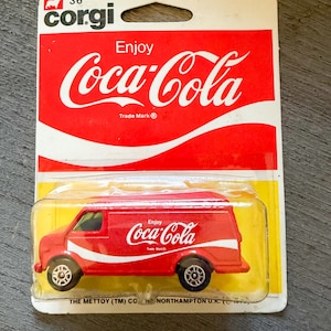 May include: A red Corgi Coca-Cola delivery van toy in its original packaging. The van has the Coca-Cola logo and a white stripe. The packaging is red and white with the Coca-Cola logo and the Corgi logo.