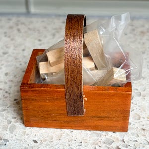 May include: A small, square wooden basket with a dark brown handle. The basket is stained orange and contains small, rectangular wooden blocks inside a clear plastic bag. The handle is made of a darker, textured wood.