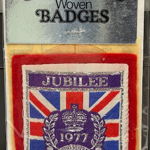 May include: A vintage woven badge from Sampson's, featuring a Union Jack design with the text "JUBILEE 1977 THE QUEEN'S SILVER JUBILEE." The badge is set on a red felt background, with a silver border.