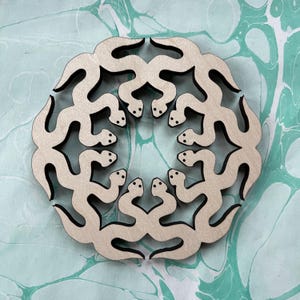 May include: A wooden coaster with a repeating pattern of stylized snakes in a circular design. The snakes are light-coloured with black outlines and small dot eyes. The coaster is on a teal and white marbled surface.