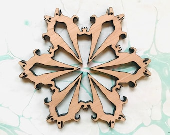 May include: A wooden snowflake ornament with a light brown finish and black detailing. The snowflake has a six-pointed design with stylized animal shapes at the ends of each point. The ornament is placed on a white and light blue marbled surface.