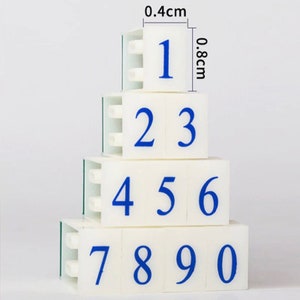 May include: A set of ten white plastic number stamps with blue numbers. The stamps are arranged in a pyramid shape, with the number 1 at the top and the numbers 7, 8, 9, and 0 at the bottom. The stamps are 0.4 cm wide and 0.8 cm tall.
