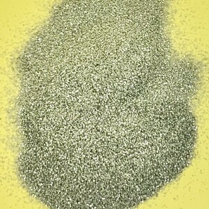 May include: A pile of fine, silver glitter on a yellow surface. The glitter particles are small and reflective, creating a shimmering effect. The glitter is densely packed, forming a textured surface.