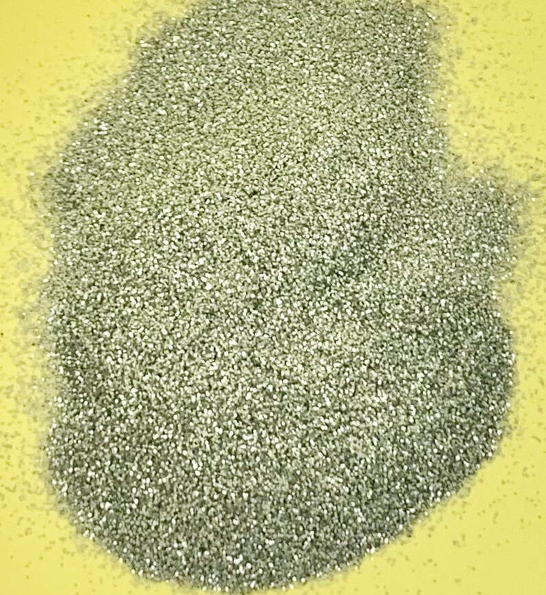 May include: A pile of fine, silver glitter on a yellow surface. The glitter particles are small and reflective, creating a shimmering effect. The glitter is densely packed, forming a textured surface.