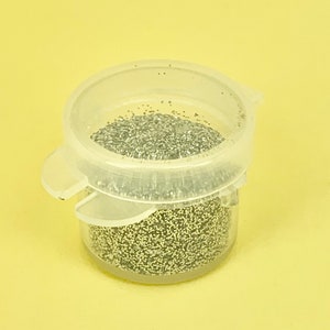 May include: A clear plastic container filled with silver glitter. The container has a hinged lid and is set against a yellow background. The glitter appears to be fine and evenly distributed within the container.