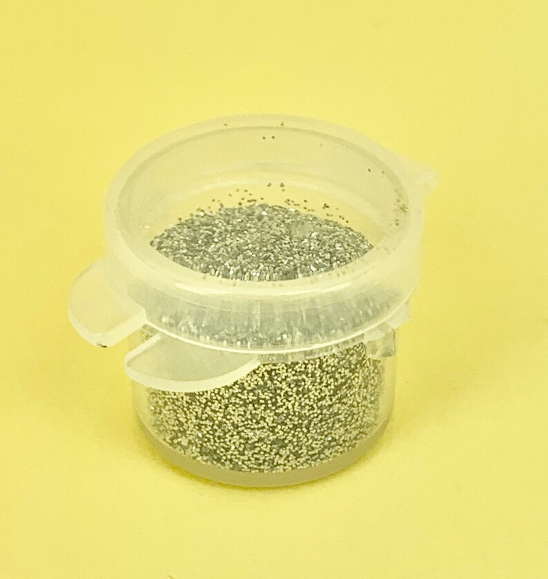 May include: A clear plastic container filled with silver glitter. The container has a hinged lid and is set against a yellow background. The glitter appears to be fine and evenly distributed within the container.