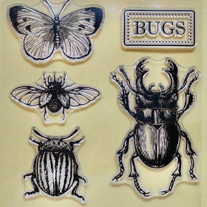 Puede incluir: Un conjunto de cinco sellos de goma transparente con ilustraciones en blanco y negro de insectos. Los sellos incluyen una mariposa, un escarabajo, un ciervo volante y una mariquita. El conjunto también incluye un sello con la palabra "BUGS" en una fuente decorativa.