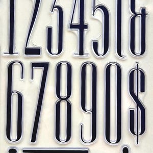 May include: A set of clear photopolymer stamps featuring the numbers 1 through 0, a dollar sign, and punctuation marks including a semicolon, a period, and quotation marks. Each character is rendered in a tall, slender, black serif typeface.