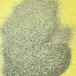 May include: A pile of silver glitter, with a reflective, textured surface. The glitter is densely packed, creating a shimmering effect. The background is a solid, light yellow color.