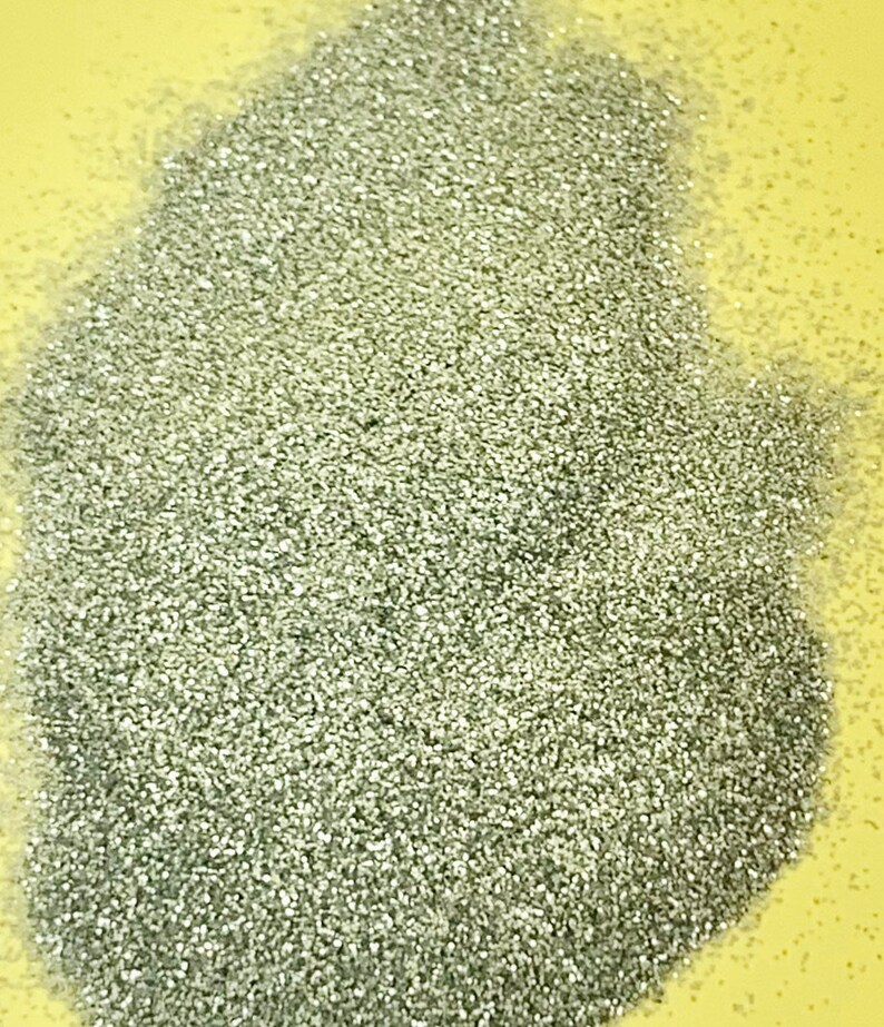 May include: A pile of silver glitter, with a reflective, textured surface. The glitter is densely packed, creating a shimmering effect. The background is a solid, light yellow color.
