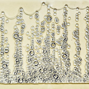 May include: Clear acrylic stamp with a bubbly design. The stamp features various sizes of clear bubbles, some with black outlines, arranged in vertical patterns against a light beige background. The top edge has a wavy, irregular shape.