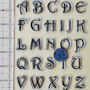 May include: A set of 26 clear alphabet stamps with black lettering. The letters are in a decorative script font. The stamps are arranged in alphabetical order.