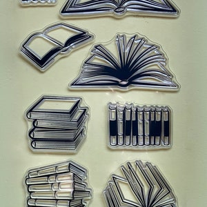 May include: A set of eight clear stamps featuring different designs of books. The stamps are arranged in two rows of four. The top row features a book with the pages open, a book with the pages closed, a book with the pages fanned out, and a book with the pages closed and the spine visible. The bottom row features a stack of books, a stack of books with the spines visible, a book with the pages fanned out, and a book with the pages closed and the spine visible. The word "Book" is printed on one of the stamps.