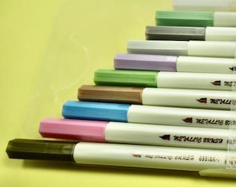 May include: A set of ten metallic brush pens with a variety of colors, including silver, gold, green, purple, and pink. The pens are arranged in a fan shape and have the text "Metallic Brush" printed on the side.