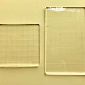 May include: Two clear acrylic stamping blocks. One is square, the other is rectangular. Both blocks have a grid pattern etched into the surface. The blocks are designed for crafting and stamping projects.