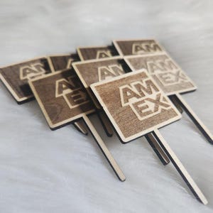 May include: A collection of wooden square toppers with the letters "AM EX" etched into the surface. Each topper has a thin, rectangular stick attached to the bottom. The wood has a natural, light brown colour.