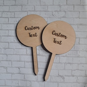 May include: Two round wooden signs with sticks attached to the back. The signs are blank and have the text "Custom Text" written on them.