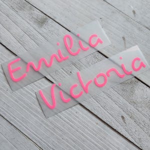 May include: Two rectangular adhesive vinyl decals with a matte translucent backing. The top decal features the name Emilia in bright pink cursive lettering, and the bottom decal features the name Victoria in the same bright pink cursive font style.