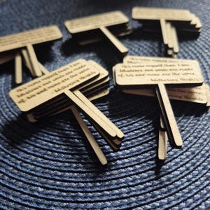May include: A collection of light brown wooden plant markers, each with a T-shape and the quote "He's more myself than I am..." from Wuthering Heights. The markers are stacked and arranged on a blue surface.