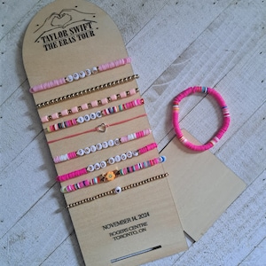 May include: A set of 8 pink and white beaded bracelets with different charms and lettering, including "Lover", "1989", "Swiftie", and "Bad Blood". The bracelets are displayed on a wooden board with the text "Taylor Swift The Eras Tour" and "November 14, 2024 Rogers Centre Toronto, ON".