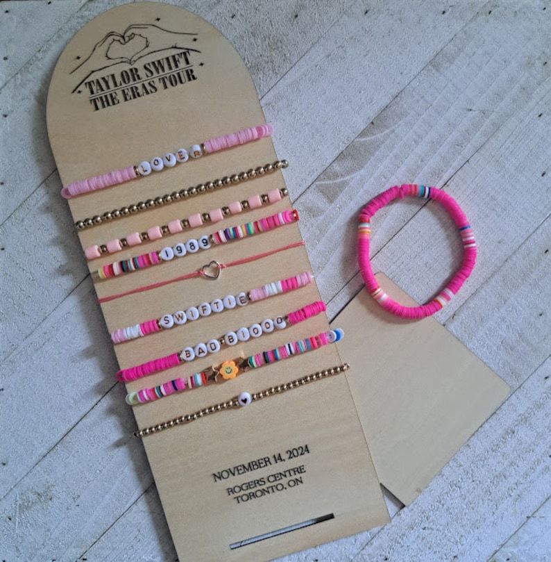 May include: A set of 8 pink and white beaded bracelets with different charms and lettering, including "Lover", "1989", "Swiftie", and "Bad Blood". The bracelets are displayed on a wooden board with the text "Taylor Swift The Eras Tour" and "November 14, 2024 Rogers Centre Toronto, ON".