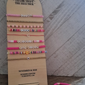 May include: A wooden display board with the text "TAYLOR SWIFT THE ERAS TOUR" and the date "NOVEMBER 14, 2024". The board has a series of colorful beaded bracelets with different words and designs, including "Lover", "1989", "Speak Now", and "Reputation".