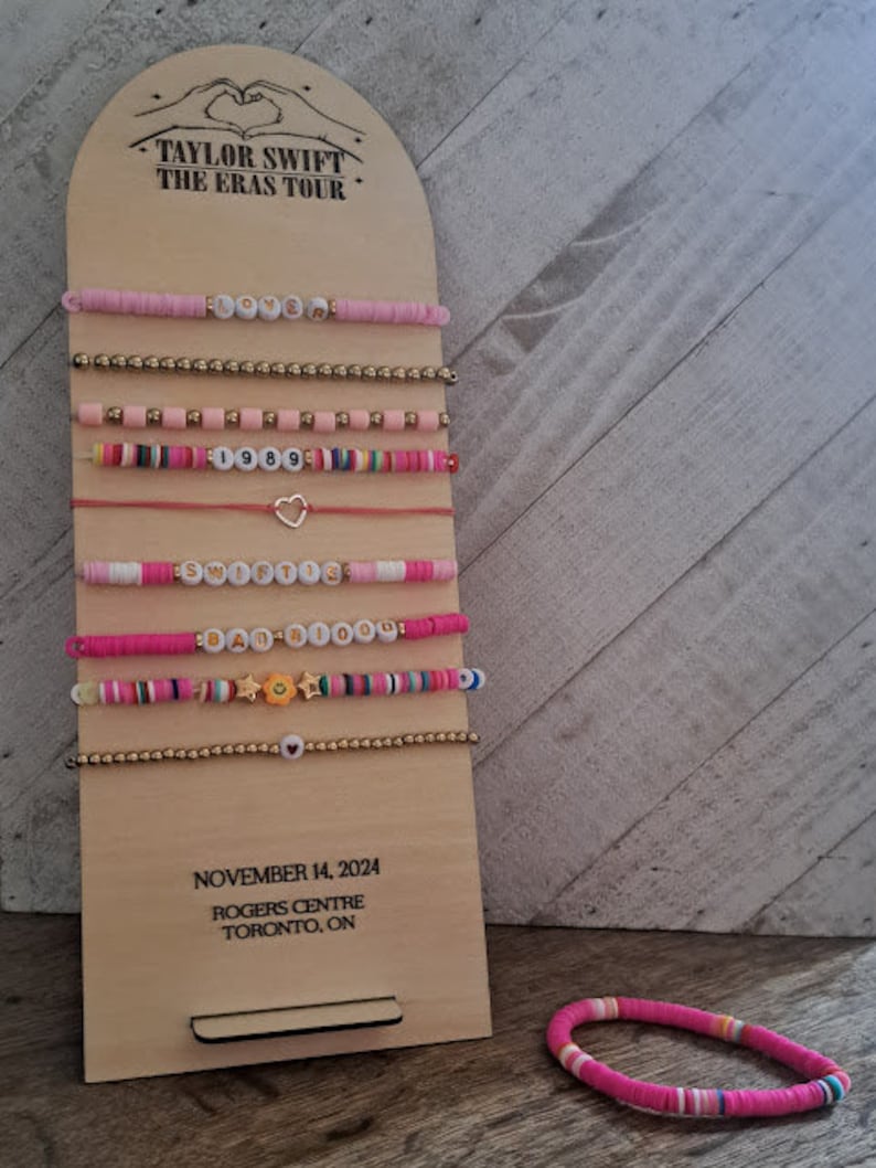 May include: A wooden display board with the text "TAYLOR SWIFT THE ERAS TOUR" and the date "NOVEMBER 14, 2024". The board has a series of colorful beaded bracelets with different words and designs, including "Lover", "1989", "Speak Now", and "Reputation".