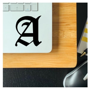 May include: A black letter A in a gothic font style is on a white laptop. The laptop is on a wooden surface.