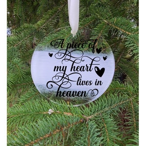 Peut inclure: Une boule de Noël en verre transparent avec un ruban blanc suspendu au sommet. La boule est ornée d'une inscription noire qui dit "A piece of my heart lives in heaven" avec un symbole de cœur.
