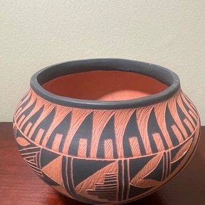 Puede incluir: Un cuenco de cerámica redondo, hecho a mano, con una abertura ancha. El cuenco presenta un borde negro y está adornado con un patrón geométrico repetitivo en negro y terracota. El interior del cuenco es de terracota.