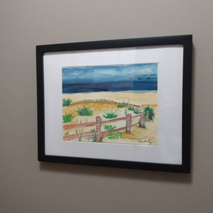 Op de afbeelding: Een aquarel schilderij van een strandscène met een houten hek, groen gras en een blauwe lucht met vogels die eroverheen vliegen. Het schilderij is ingelijst in zwart.