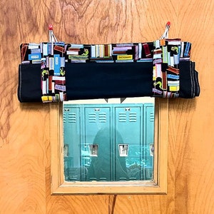 May include: A black fabric doorstop with a colorful book print design hangs on a wooden door. The doorstop has a pocket for holding items. The doorstop is hanging over a window that shows a row of blue lockers.