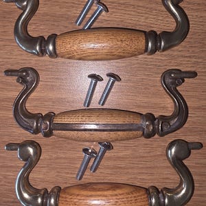 May include: Three drawer pulls featuring a central oval-shaped light brown wood grain handle flanked by curved metallic brass-toned arms. Each pull is accompanied by two silver-colored metal mounting screws with rounded heads for installation.