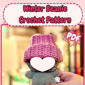 K&ouml;nnte beinhalten: Ein graues Pl&uuml;schtier mit einer rosa geh&auml;kelten M&uuml;tze. Das Bild enth&auml;lt den Text "Winter Beanie Crochet Pattern" und "PDF". Der Text "By: ChainStitchery" befindet sich unten im Bild.