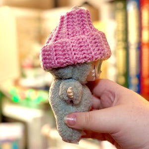 K&ouml;nnte beinhalten: Eine kleine Pl&uuml;schpuppe mit grauem K&ouml;rper und einem geh&auml;kelten rosa Hut. Die Puppe hat ein einfaches Design mit sichtbaren N&auml;hten und einem kleinen Gesicht. Der Hut ist ein leuchtendes Rosa, das mit der ged&auml;mpften grauen Farbe der Puppe kontrastiert. Die Puppe wird in einer Hand gehalten.