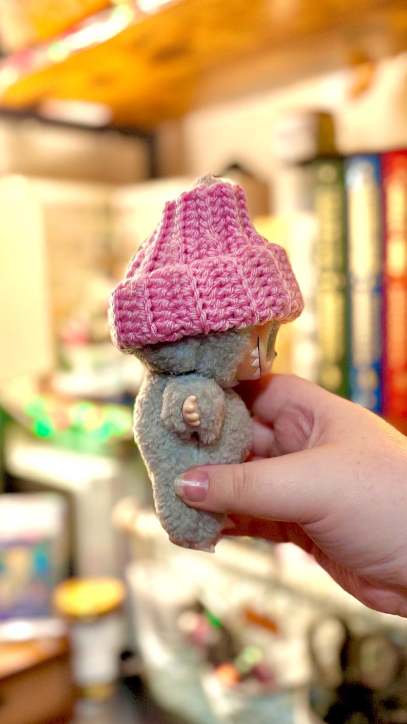 K&ouml;nnte beinhalten: Eine kleine Pl&uuml;schpuppe mit grauem K&ouml;rper und einem geh&auml;kelten rosa Hut. Die Puppe hat ein einfaches Design mit sichtbaren N&auml;hten und einem kleinen Gesicht. Der Hut ist ein leuchtendes Rosa, das mit der ged&auml;mpften grauen Farbe der Puppe kontrastiert. Die Puppe wird in einer Hand gehalten.
