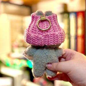 K&ouml;nnte beinhalten: Ein handgefertigtes Pl&uuml;schtier in Form eines kleinen Tieres, das einen rosa geh&auml;kelten Hut mit einem goldfarbenen Ring tr&auml;gt. Das Spielzeug ist grau und hat eine flauschige Textur. Der Hut hat ein Rippendesign.