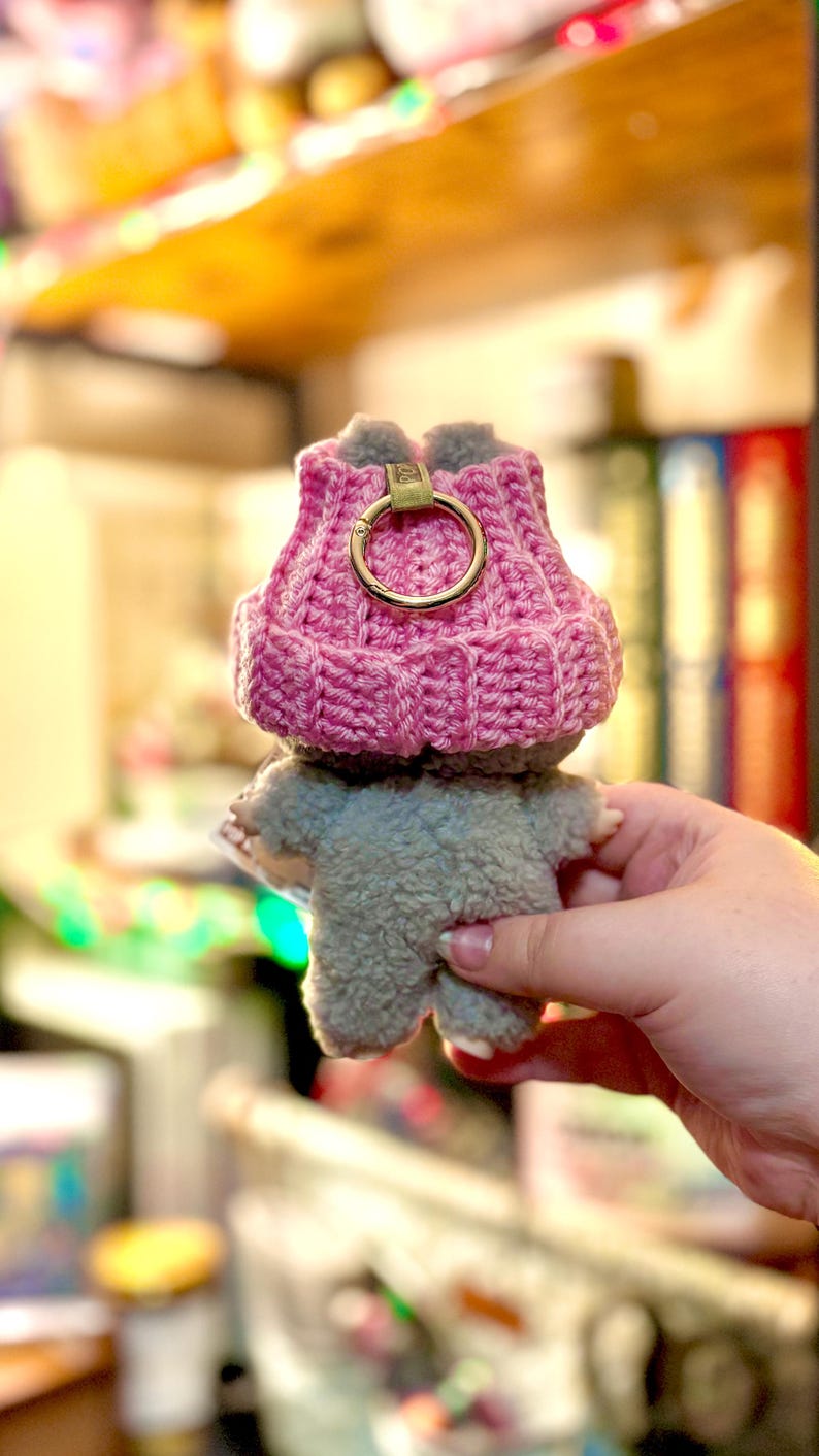 K&ouml;nnte beinhalten: Ein handgefertigtes Pl&uuml;schtier in Form eines kleinen Tieres, das einen rosa geh&auml;kelten Hut mit einem goldfarbenen Ring tr&auml;gt. Das Spielzeug ist grau und hat eine flauschige Textur. Der Hut hat ein Rippendesign.