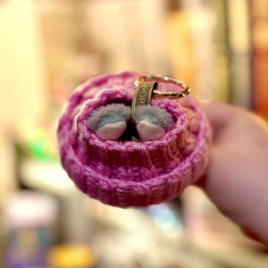 K&ouml;nnte beinhalten: Nahaufnahme einer kleinen, rosa gestrickten M&uuml;tze mit einem goldfarbenen Ring und einem kleinen Anh&auml;nger mit der Aufschrift "POP". Die M&uuml;tze enth&auml;lt zwei winzige, grau-wei&szlig;e Schuhe. Der Hintergrund ist unscharf.