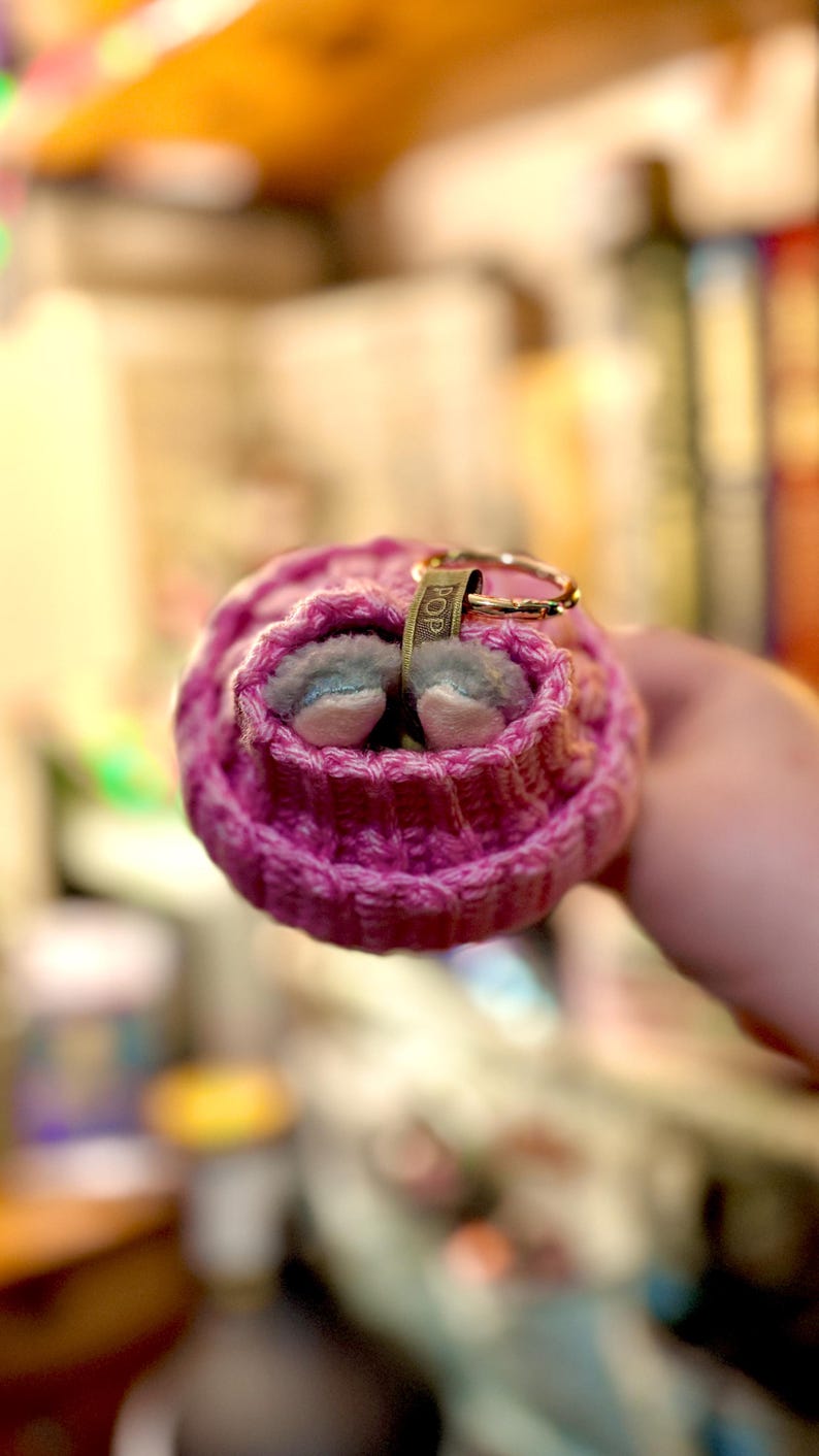 K&ouml;nnte beinhalten: Nahaufnahme einer kleinen, rosa gestrickten M&uuml;tze mit einem goldfarbenen Ring und einem kleinen Anh&auml;nger mit der Aufschrift "POP". Die M&uuml;tze enth&auml;lt zwei winzige, grau-wei&szlig;e Schuhe. Der Hintergrund ist unscharf.