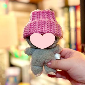 K&ouml;nnte beinhalten: Ein kleines, graues Pl&uuml;schtier mit einer rosa Strickm&uuml;tze. Das Spielzeug hat ein herzf&ouml;rmiges Gesicht und wird in der Hand einer Person gehalten. Die M&uuml;tze ist leuchtend rosa und hat eine gerippte Textur. Der K&ouml;rper des Spielzeugs ist aus weichem, flauschigem Material.