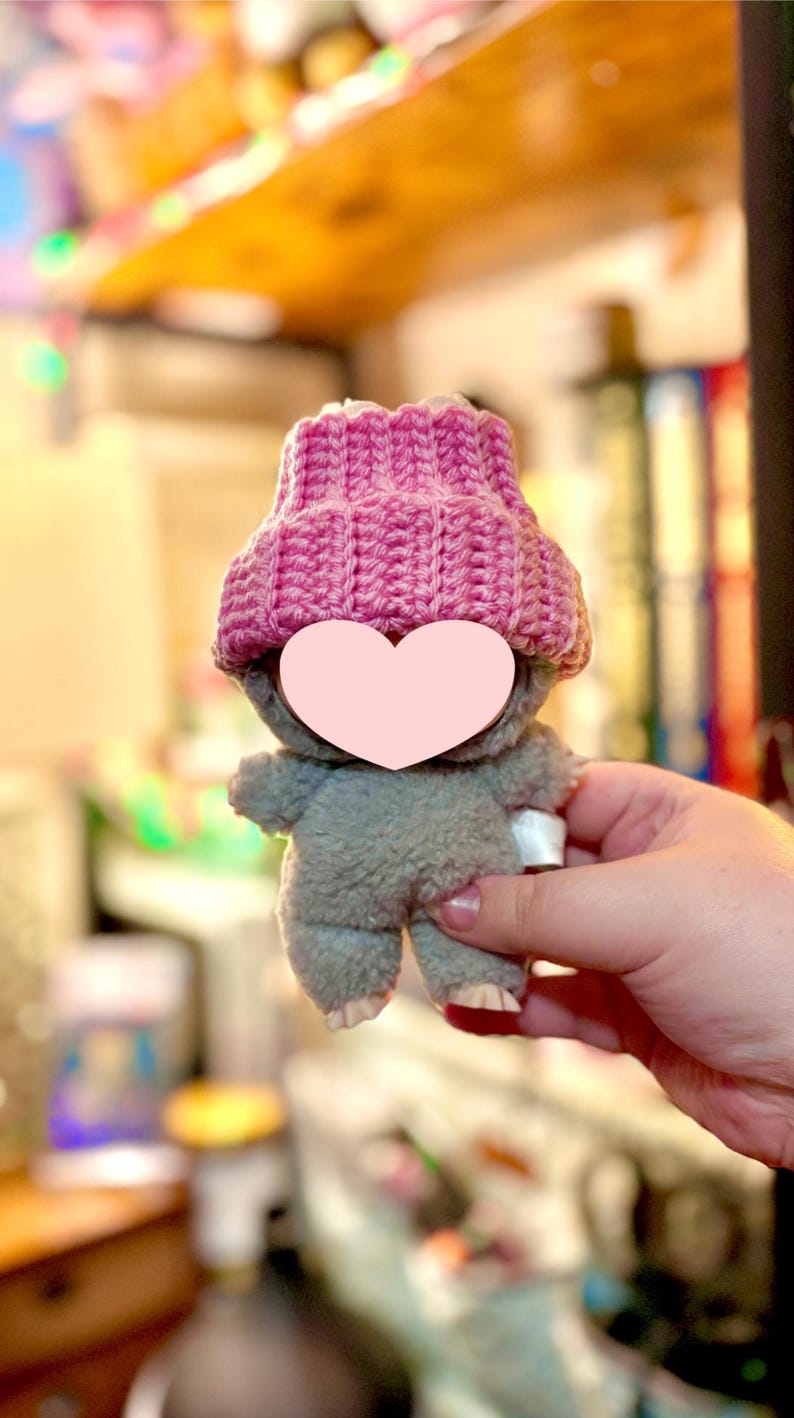 K&ouml;nnte beinhalten: Ein kleines, graues Pl&uuml;schtier mit einer rosa Strickm&uuml;tze. Das Spielzeug hat ein herzf&ouml;rmiges Gesicht und wird in der Hand einer Person gehalten. Die M&uuml;tze ist leuchtend rosa und hat eine gerippte Textur. Der K&ouml;rper des Spielzeugs ist aus weichem, flauschigem Material.